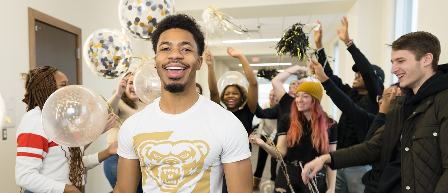 A young man smiling with a crowd behind him, holding black and gold balloons and ribbons.