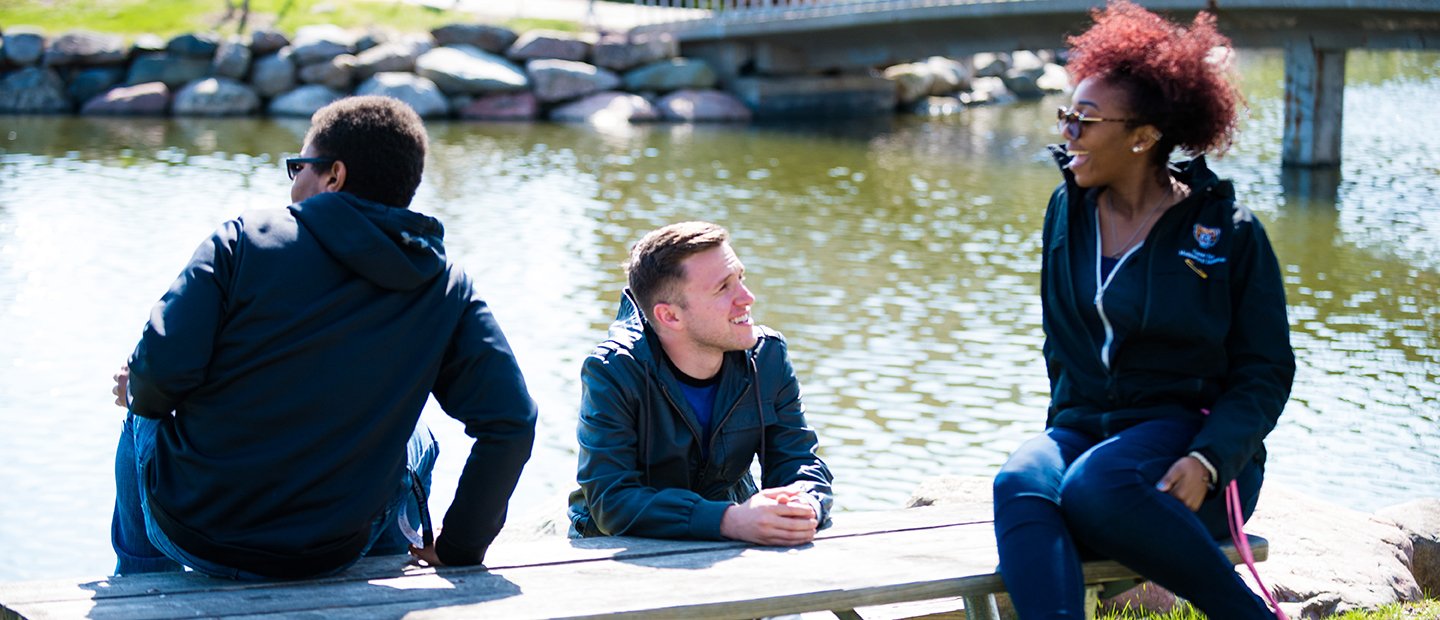 Three students sitting on a picnic table in front of Bear Lake at Oakland University.