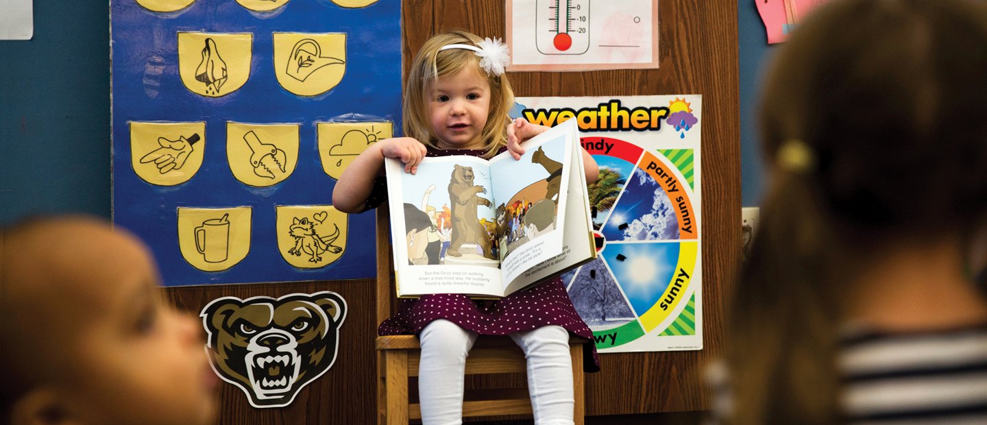 Young girl seated at the front of a classroom, showing her classmates a book.