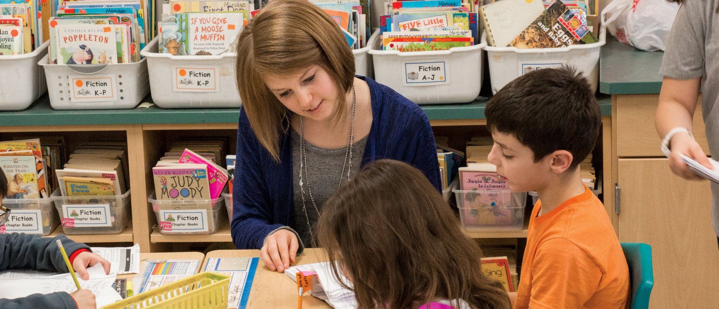 A female teacher seated with young students, reading books.