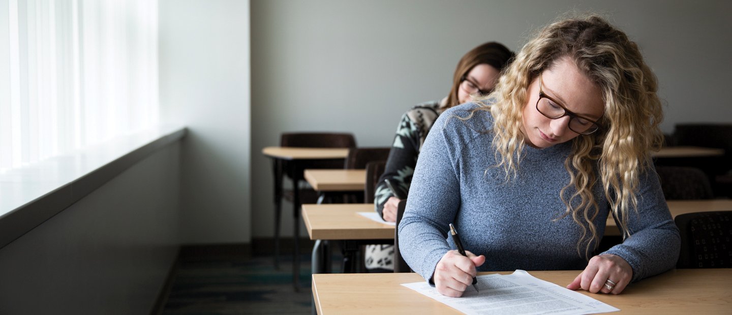 Two female students seated at desks, writing on documents.