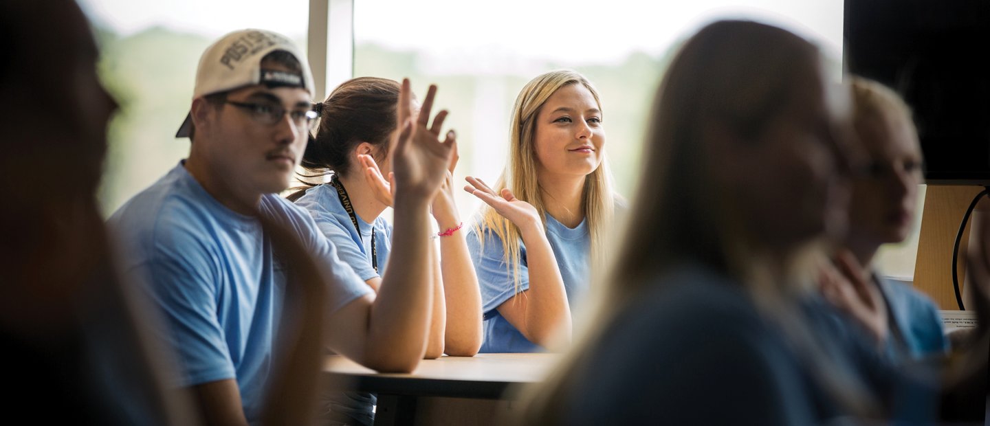 Young adults in blue t-shirts, seated at desks, raising their hands.