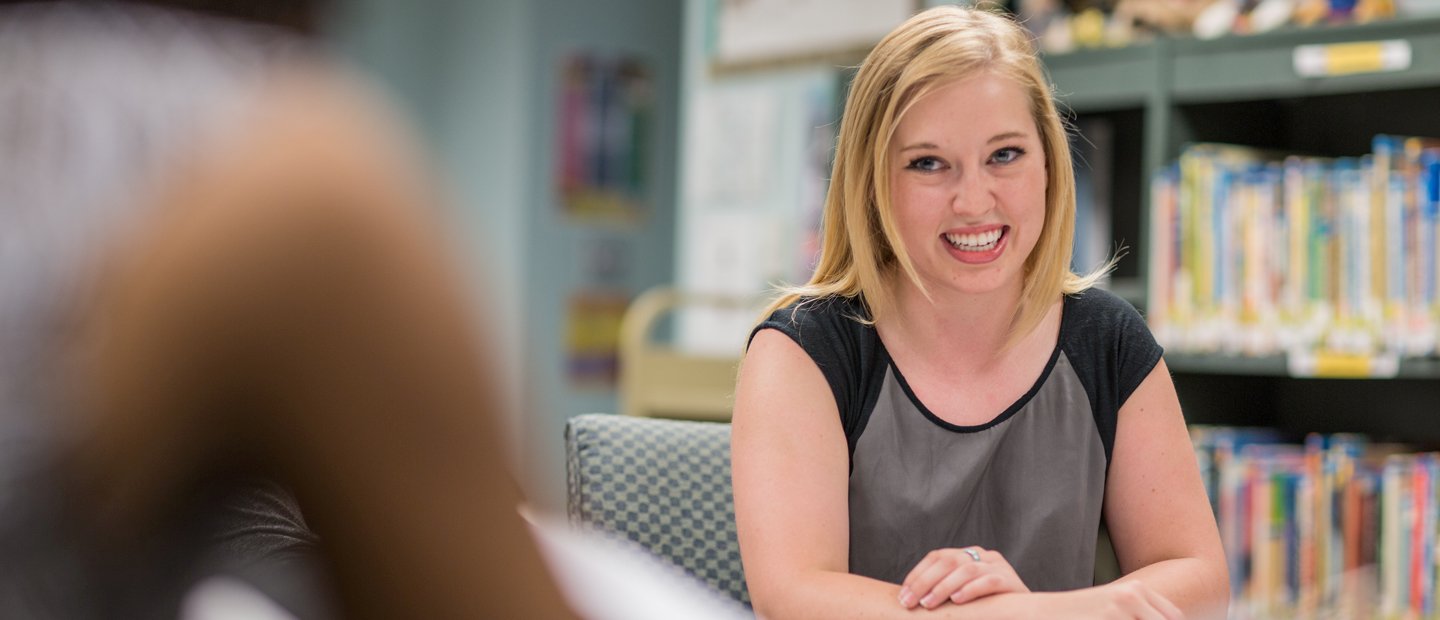 A young woman seated at a table in a library, smiling.