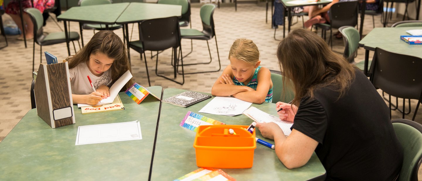 Two young girls sitting with a woman at a green table, writing and reading.