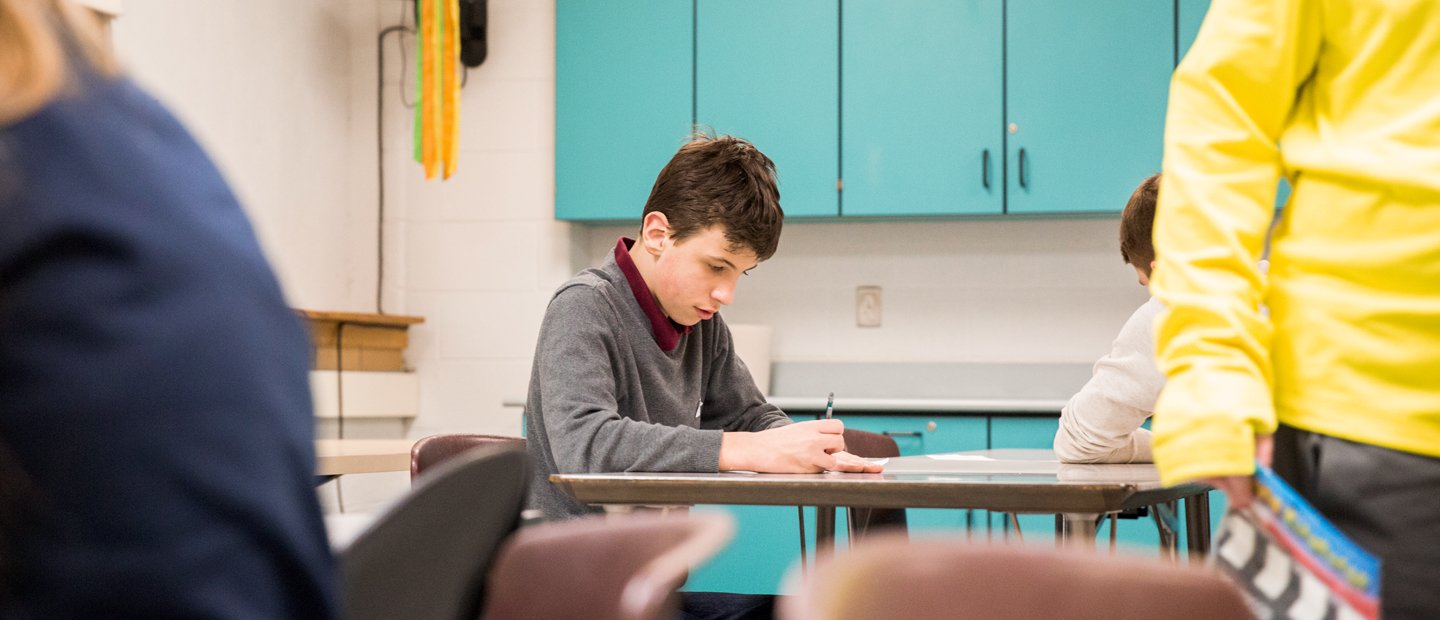 A boy seated at a table in a classroom, writing.