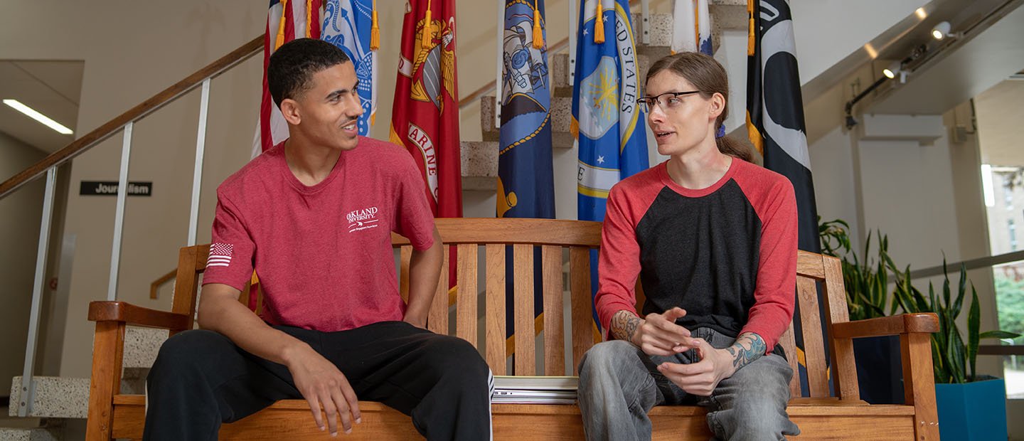 Two young men sit on a bench talking. There are a flags behind them.