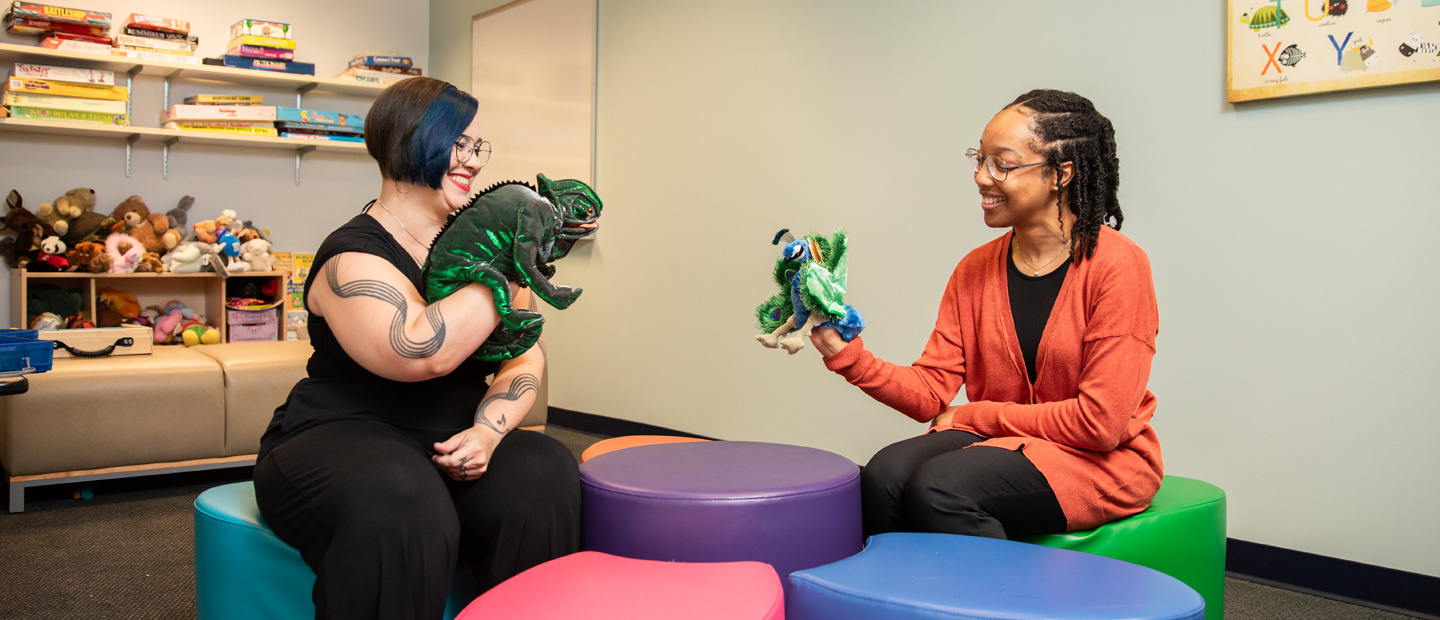 Two female students sitting across from each other smiling with puppets on their hands