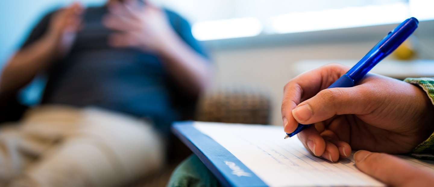 A hand holding a blue pen, writing on a notepad. Another person gesturing with his hands in the background.