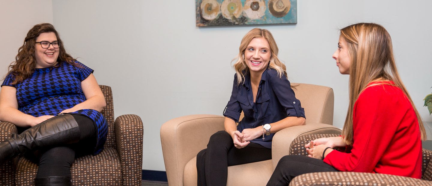 Three women seated in arm chairs, facing each other, smiling.