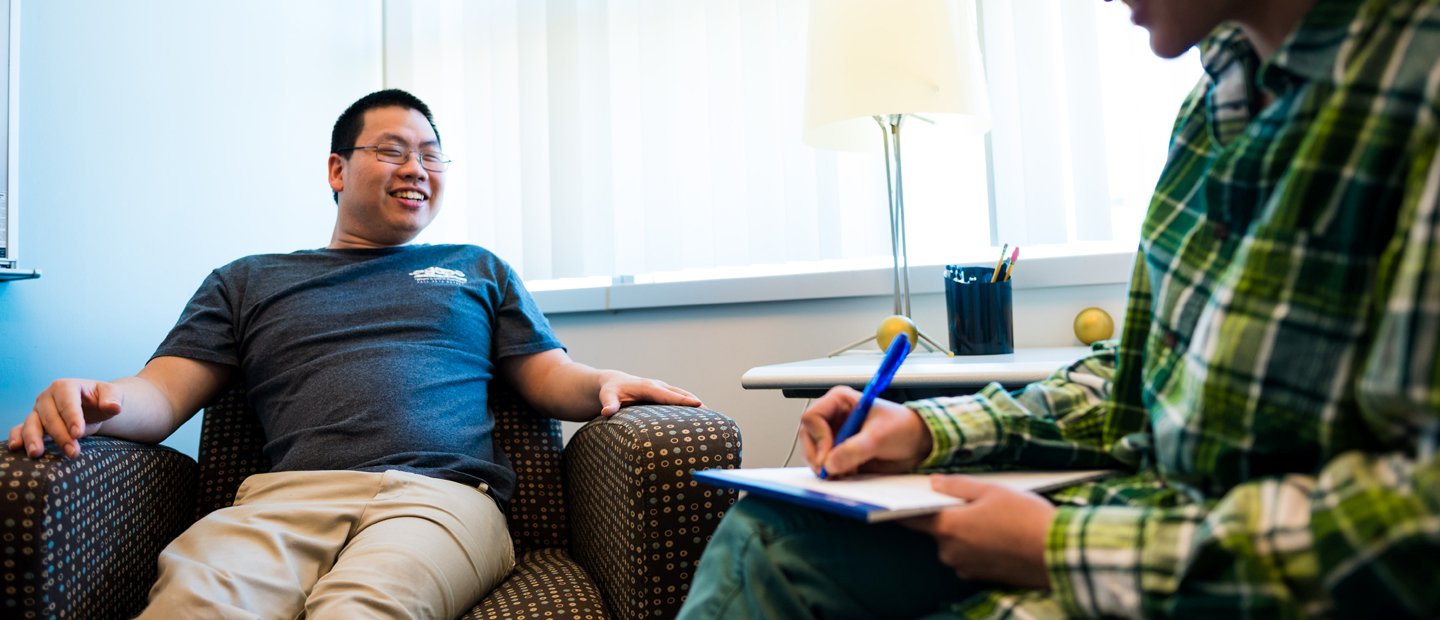 A young man seated in an arm chair smiling, facing another person who is writing on a notepad.