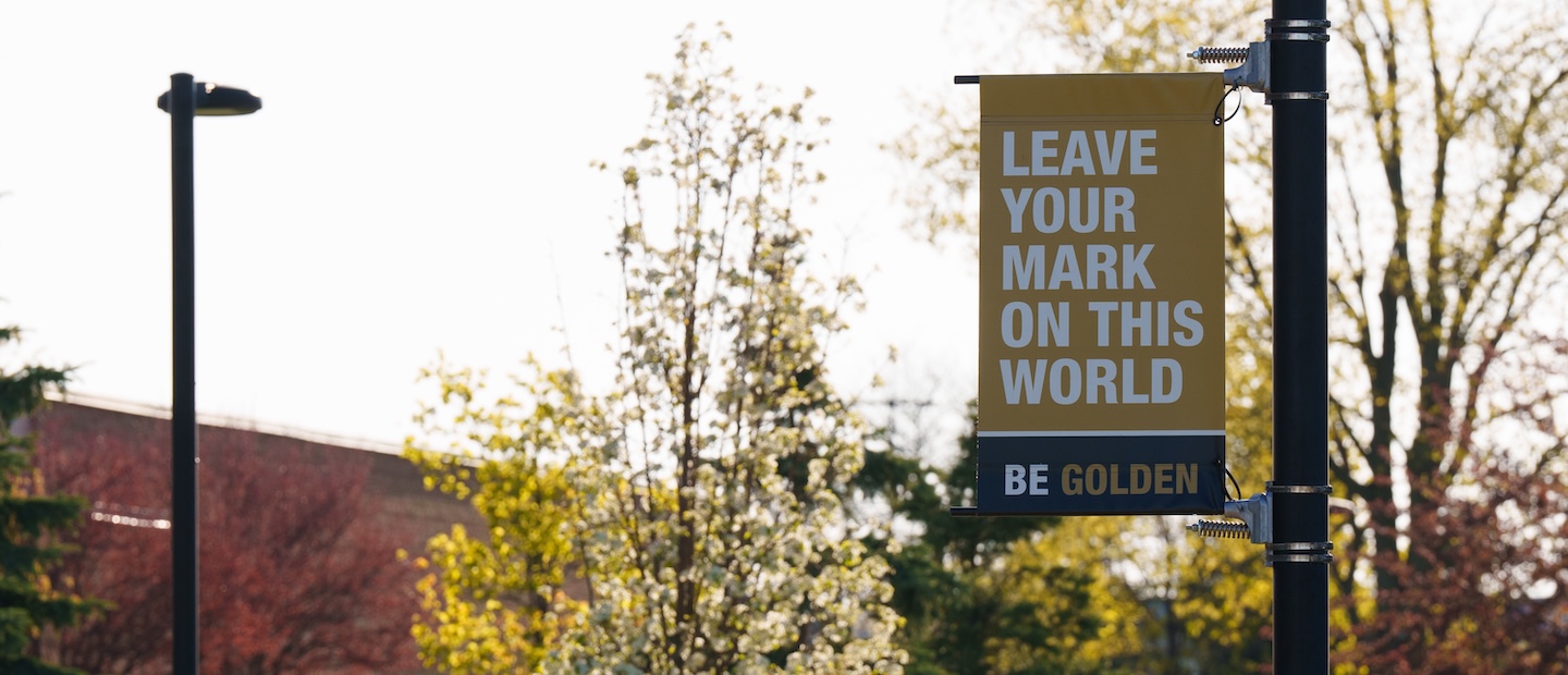 A banner that reads “Leave Your Mark on This World, Be Golden” on a pole in front of trees on Oakland University’s campus.