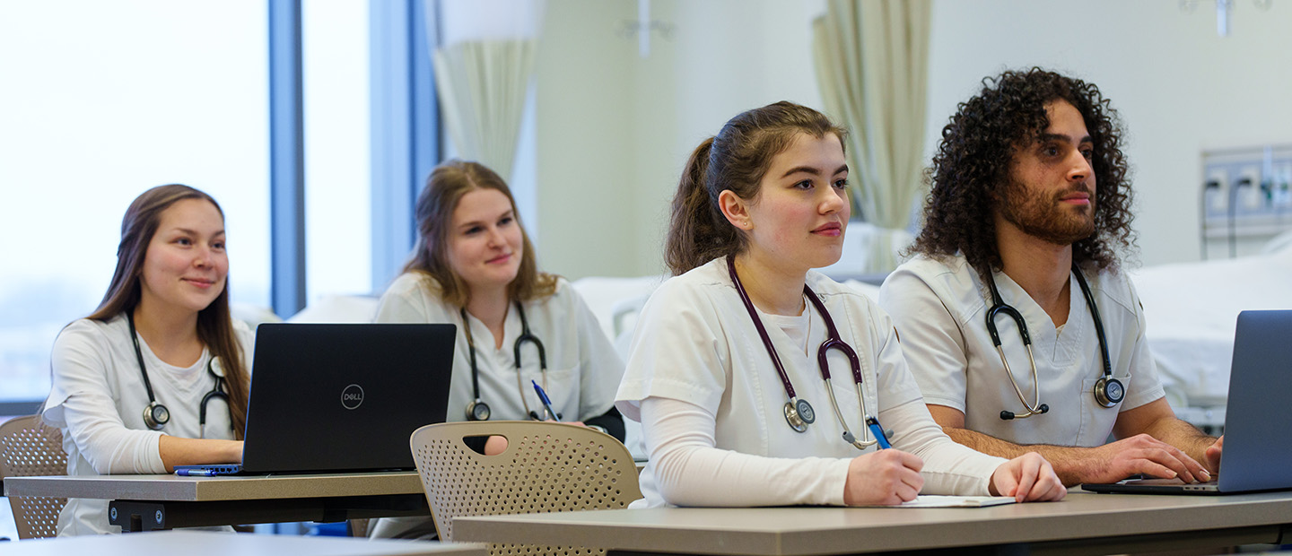 four students in white lab coats seated at a desk with stethoscopes