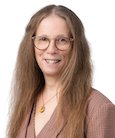 Headshot: long haired woman with glasses in med. brown top smiling at the camera.