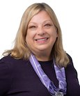 Headshot: Blonde woman in Black top with purple accent smiling at the camera