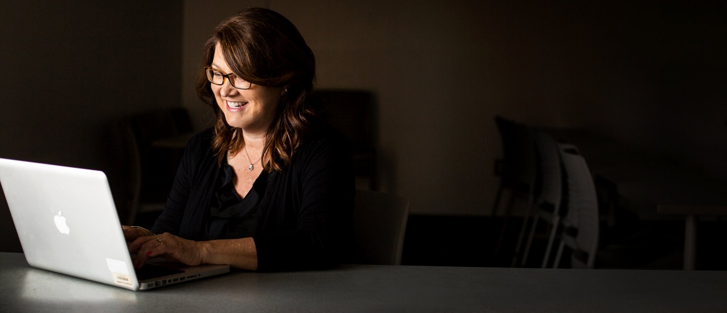 woman seated at a table with an open laptop in a dark room