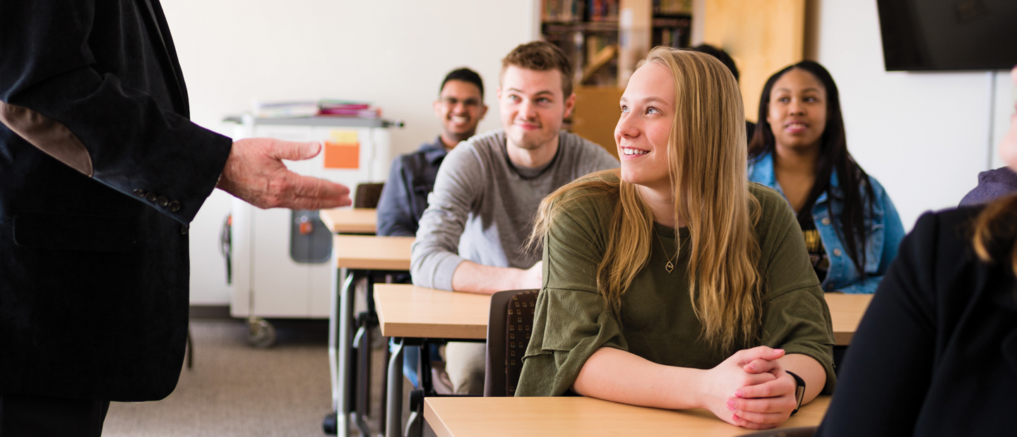 students in a classroom seated at desks, all watching a professor in front