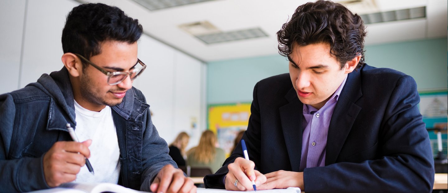 two young men seated at a desk in a classroom, writing in notebooks