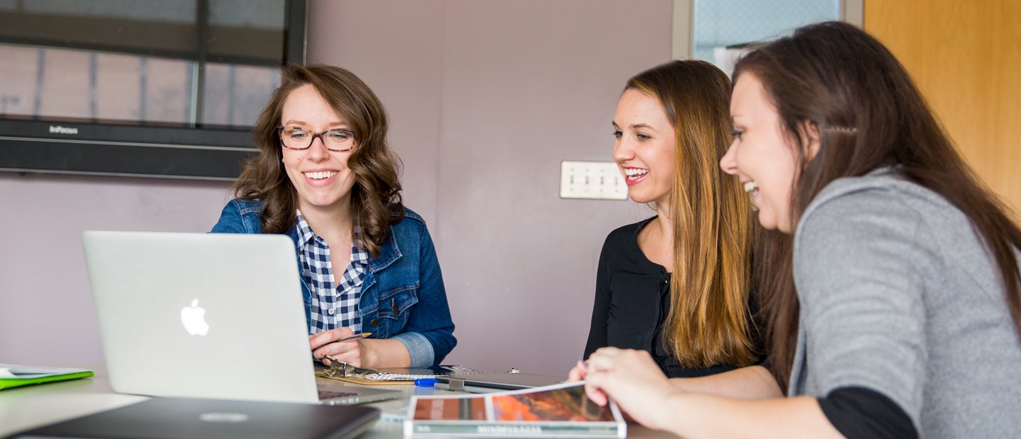 Three women seated at a table, looking at a laptop together.