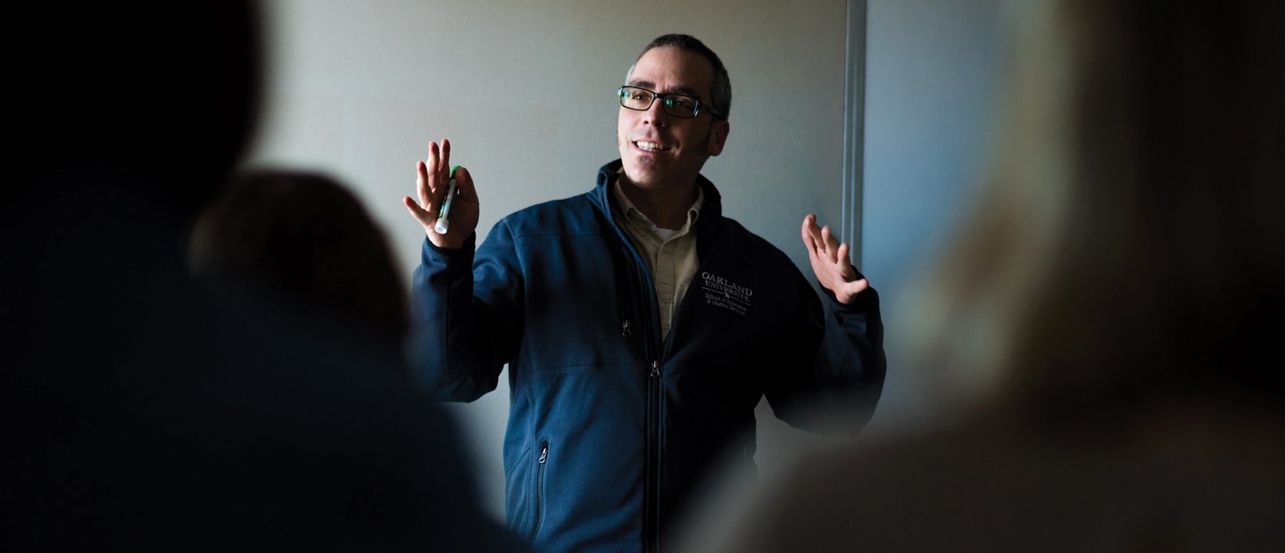 A teacher standing in front of a class, gesturing with his hands.