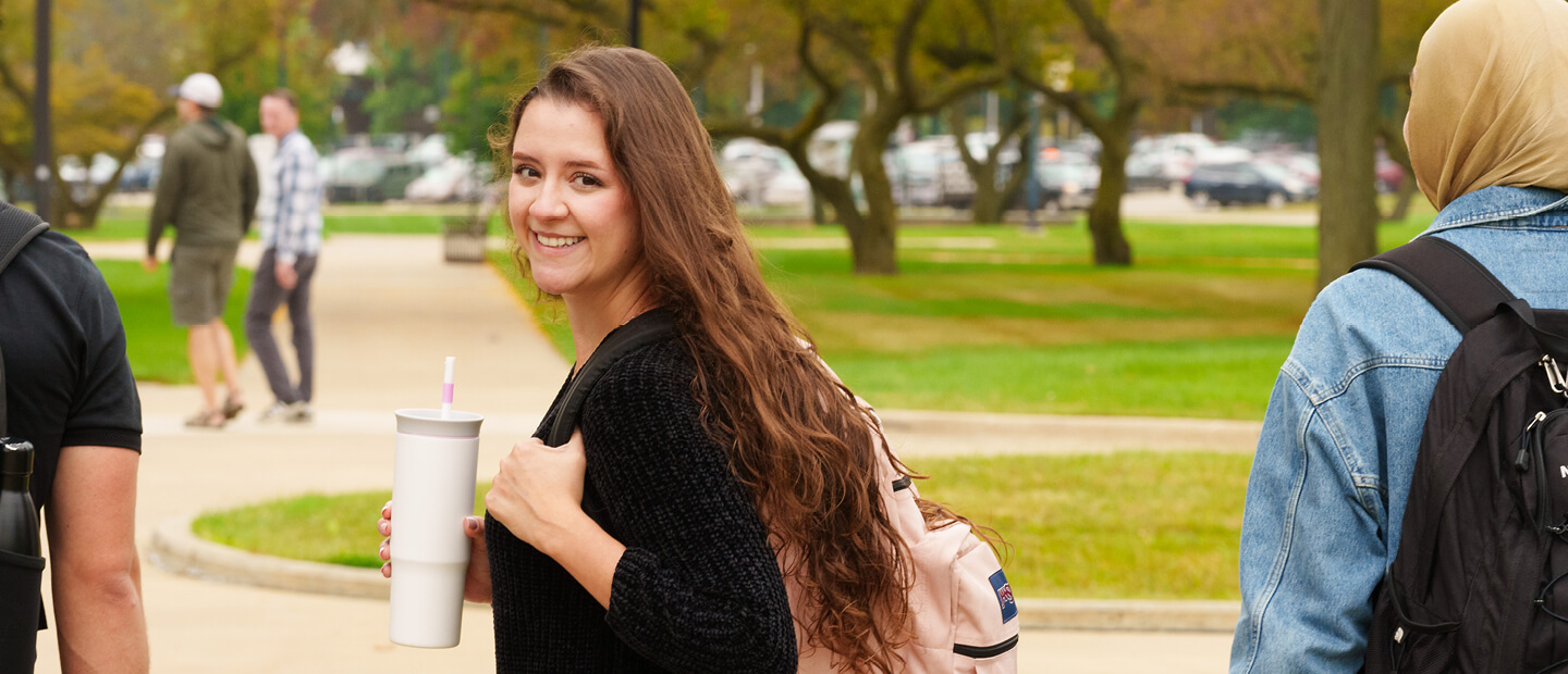Female student with long brown hair holding a mug and looking back at the camera.
